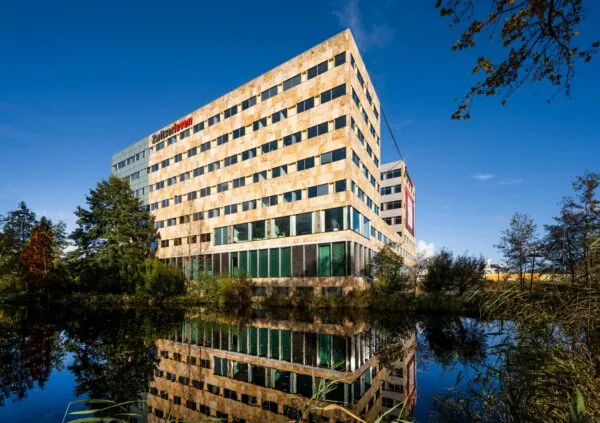 Kantoorgebouw van Zwitserleven aan de Burgemeester Rijnderslaan, weerspiegeld in het water onder een heldere blauwe lucht.